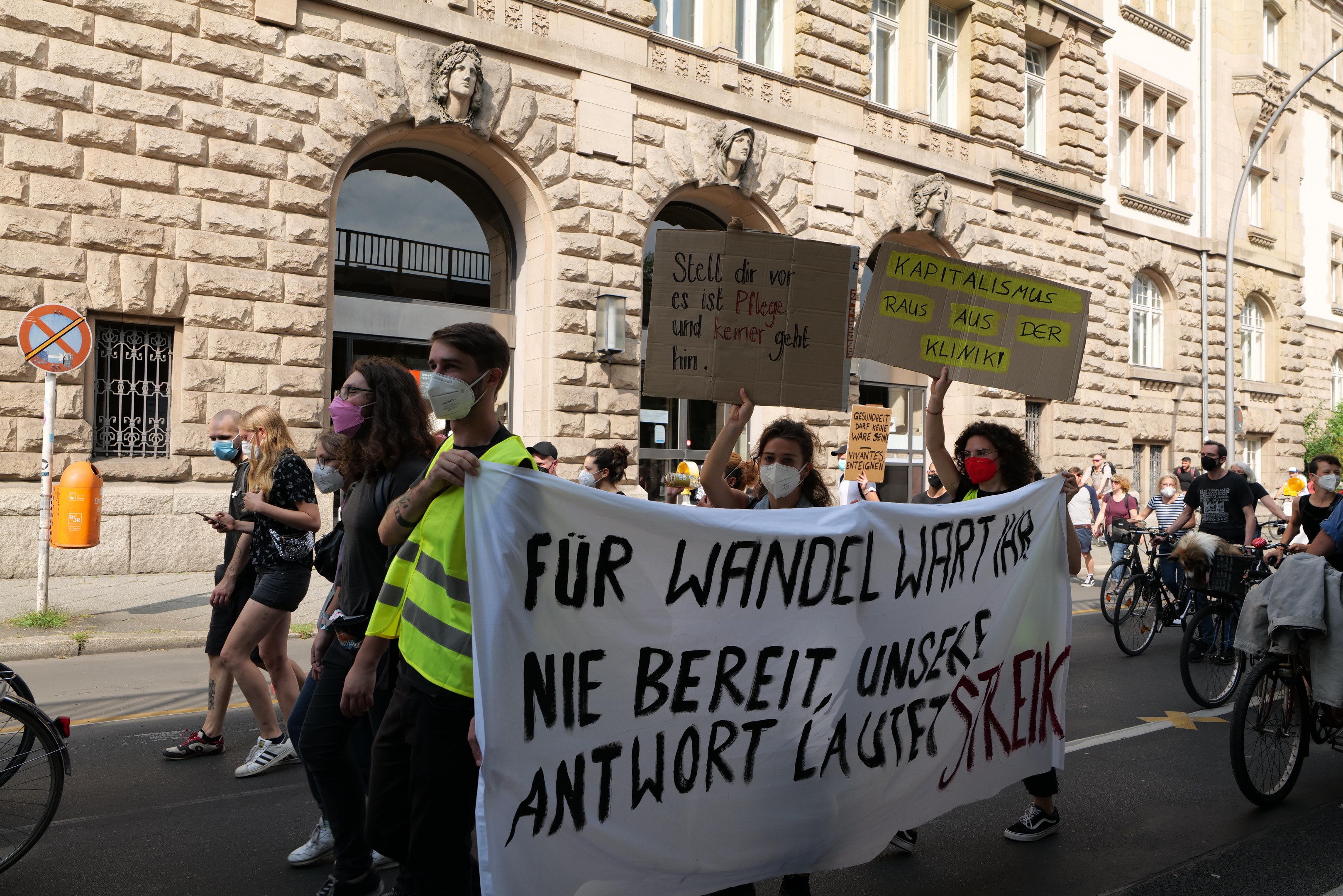 Eine Gruppe von Menschen, die eine Straße entlanggehen, einige halten Schilder und Banner, während andere Fahrräder fahren, mit einem Gebäude im Hintergrund, das Fenster, Bögen, Säulen und Skulpturen aufweist, sowie Bäume und einen Laternenpfahl während einer Protestaktion gegen die Anti-Kriegsbewegung in Berlin.