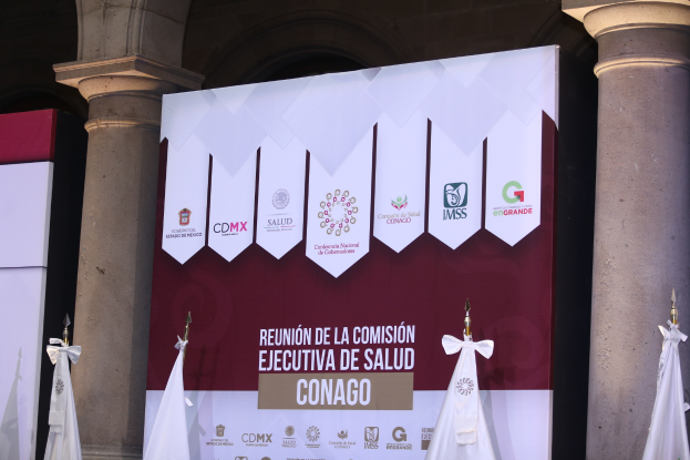 Eine Gruppe von Menschen steht vor einem Gebäude mit einer Tafel, auf der "Reunion de la Comisión Ejecutiva de Salud Conago" steht, in der Mitte. Kleidung hängt über den Säulen.