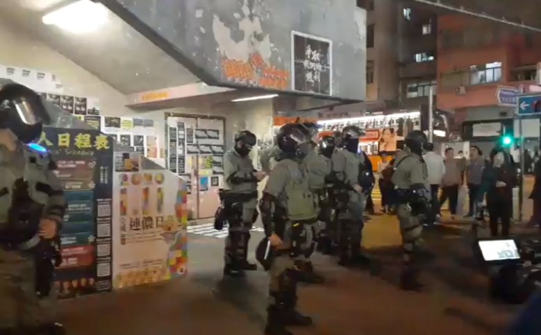 Polizisten in Schutzausrüstung stehen vor einer Menge von Helmträgern mit Waffen, mit Plakaten an Wänden und Gebäuden im Hintergrund, in einer Szene aus Hong Kong, wo die Polizei versucht, die Menge zu zerstreuen. Ein Verkehrslicht und ein Schild sind auf der rechten Seite zu sehen.