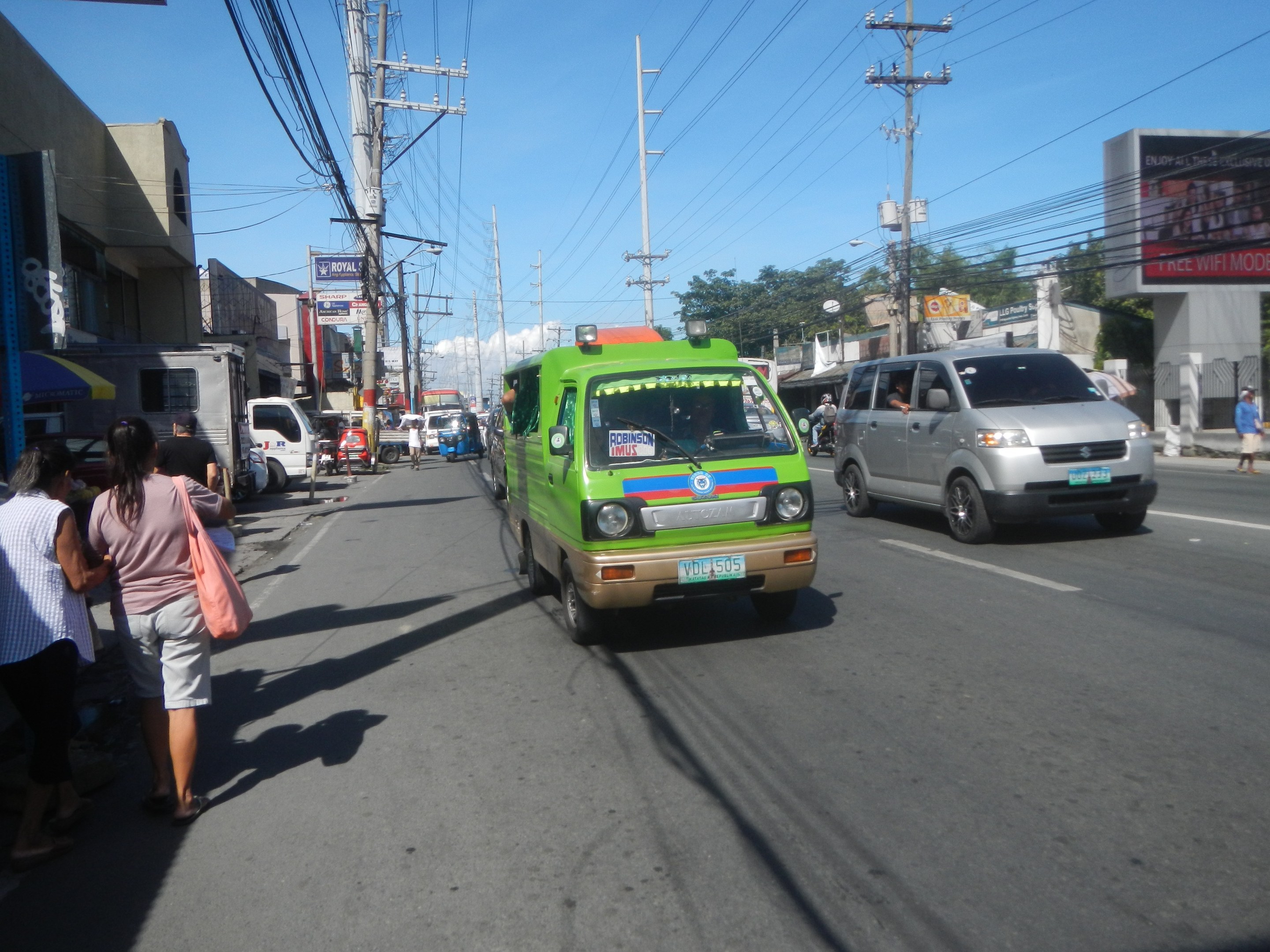 Ein grüner Lieferwagen fährt auf einer Straße mit hohen Gebäuden auf beiden Seiten, Passanten auf dem Gehweg, Strommasten mit Drähten, Bäumen im Hintergrund und einem klaren blauen Himmel.