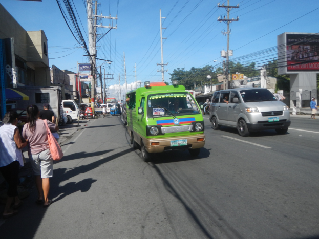 Ein grüner Lieferwagen fährt auf einer Straße mit hohen Gebäuden auf beiden Seiten, Passanten auf dem Gehweg, Strommasten mit Drähten, Bäumen im Hintergrund und einem klaren blauen Himmel.