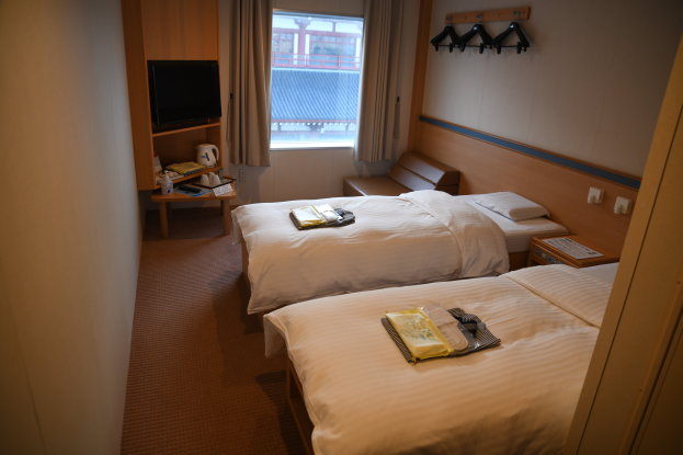 Ein Hotelzimmer mit zwei Betten, einem Wandfernseher, einem Fenster mit Vorhängen, Bügeln auf der rechten Seite und einem Tisch mit verschiedenen Gegenständen vor den Betten.