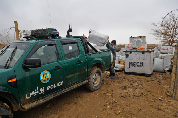 Ein Polizei-Lkw steht auf der Seite eines Schotterwegs neben einem Zaun, zwei Personen sind in der Nähe, eine hält eine Tasche, und diverse Gegenstände sind im Hintergrund zu sehen.