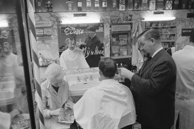 Schwarz-weißes Foto eines Mannes, der einer Frau in einem Barbiersalon die Haare schneidet, mit einem Waschbecken, einem Spiegel, Regalen und einer Glastür im Hintergrund.