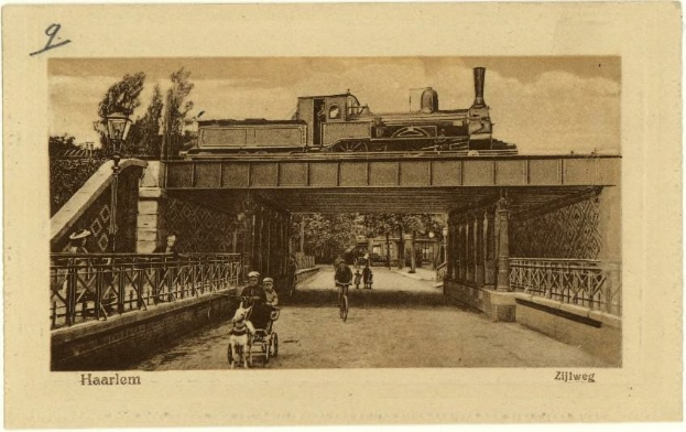 Schwarz-weiß-Foto eines Zuges, der über eine Brücke fährt, mit Menschen auf Fahrrädern und einer Person in einem Kinderwagen, Bäume im Hintergrund und Text unten.