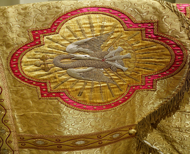 Nahaufnahme eines Priestergewandes mit goldenen und roten Stickereien und detaillierten Mustern, vor einer Wand im Hintergrund.