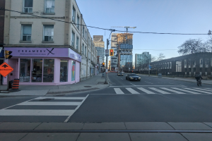 Stadtstraße mit fahrenden Autos, ein Radfahrer auf dem Gehweg, Verkehrszeichen, Straßenschilder, Gebäude, Bäume und Himmel.