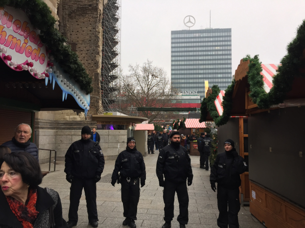Polizeibeamte in schwarzen Uniformen stehen vor einem geschmückten Weihnachtsmarkt in Berlin, mit festlichen Dekorationen, Gebäuden, Bäumen und einem klaren blauen Himmel im Hintergrund.