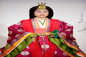 Eine Frau in einem traditionellen japanischen Kimono, das ein lebhaftes rotes und grünes Kleid mit aufwändigen Mustern zeigt, steht selbstbewusst auf einem Laufsteg, ihr Haar ist ordentlich zu einem Knoten gebunden.