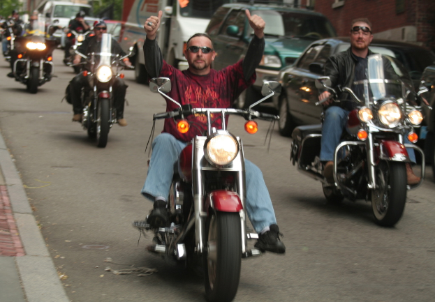 Eine Gruppe von Menschen, die auf Fahrrädern auf einer Straße fahren, wobei die Person in der Mitte ein rotes Hemd trägt und ohne Hände fährt, und eine weitere Person rechts mit einer schwarzen Jacke und einem Lachen, mit zwei Autos und einem Lieferwagen im Hintergrund.