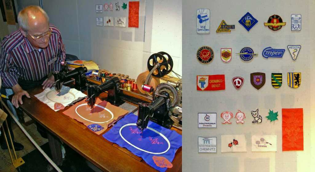 Ein Mann mit Brille steht hinter einem Tisch mit einer Platte, auf der Abzeichen, Nähmaschinen, Kleidung und verschiedene farbige Fäden zu sehen sind.