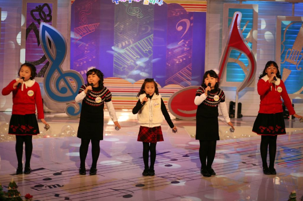 Eine Gruppe von Kindern steht auf einer Bühne, jeder hält ein Mikrofon in der Hand, mit einem bunten Hintergrund dahinter.