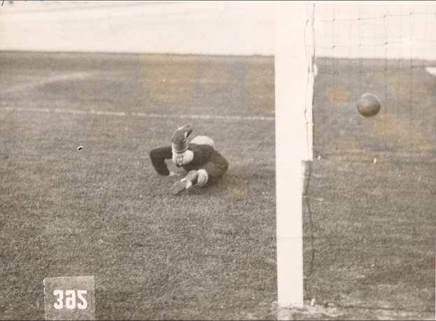 Ein Schwarz-Weiß-Foto eines Fußballspielers, der einen Ball vor einem Torpfosten mit Netz auf der rechten Seite fängt und Text unten.