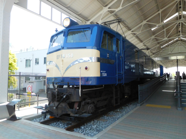 Ein blauer und weißer Zug fährt auf Schienen neben einem Bahnsteig mit einigen Menschen, Treppen auf der rechten Seite und einem Geländer auf der linken Seite, mit Bäumen, einem Gebäude und dem Himmel im Hintergrund.
