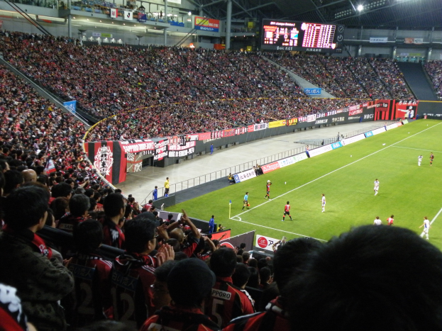 Ein Fußballspiel in einem großen Stadion mit Spielern auf dem Feld und Zuschauern auf den Tribünen, umgeben von Bannern, einem Zaun, einem Display, Metallstangen und einem Dach mit Deckenleuchten.