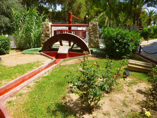 Minigolfanlage in einem Park mit Grünflächen, einer Straße mit Fußgängern, Straßenschildern und einem blauen Himmel im Hintergrund.