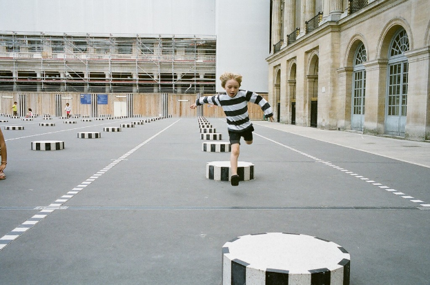 Ein Spielbereich mit vier Kindern, eines davon rennt auf dem Boden mit schwarzen und weißen Betonstufen, umgeben von einem Schulgeb├Ąude.