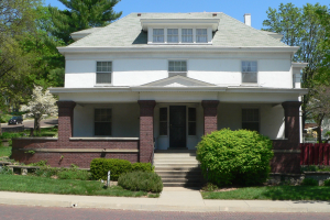 Ein weißes Haus mit einem grünen Dach und einem roten Zaun, umgeben von Pflanzen, Gras, einem Pfad, einem Auto auf der Straße, einer Gruppe von Bäumen und einem bewölkten Himmel.