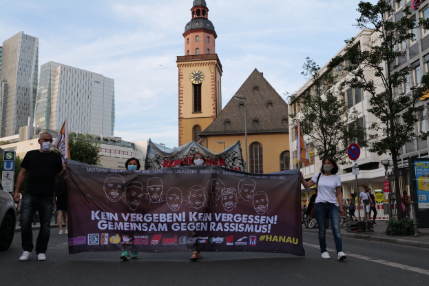 Eine Gruppe maskierter Personen marschiert auf einer Straße und hält ein Banner, daneben steht ein geparktes Auto, im Hintergrund sind Gebäude, Bäume, Schilder, Pfosten und ein Uhrenturm unter einem klaren blauen Himmel zu sehen.