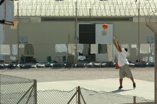 Ein Mann in sportlicher Schuhen bereit macht sich zum Basketballwurf vor einem Zaun, mit einem Basketballkorb links daneben und einem Gebäude im Hintergrund.