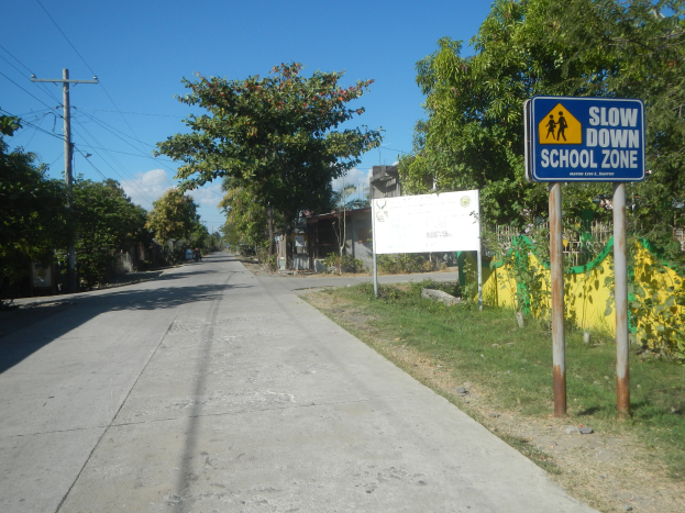 Eine Straße mit einem "Slow Down School Zone"-Schild, Häuser, Bäume und ein bewölkter Himmel, mit Strommasten und -leitungen im Hintergrund.