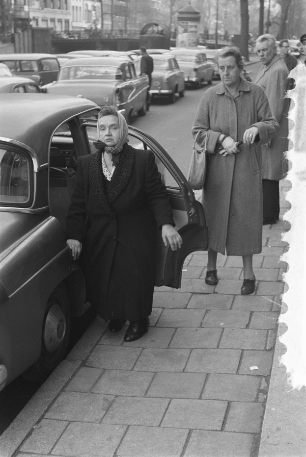 Eine Schwarz-Weiß-Szene einer Frau, die aus einem Auto auf einer Stadtstraße aussteigt, mit anderen Fahrzeugen, Passanten, Bäumen, Pfählen, Gebäuden und einem klaren Himmel im Hintergrund.