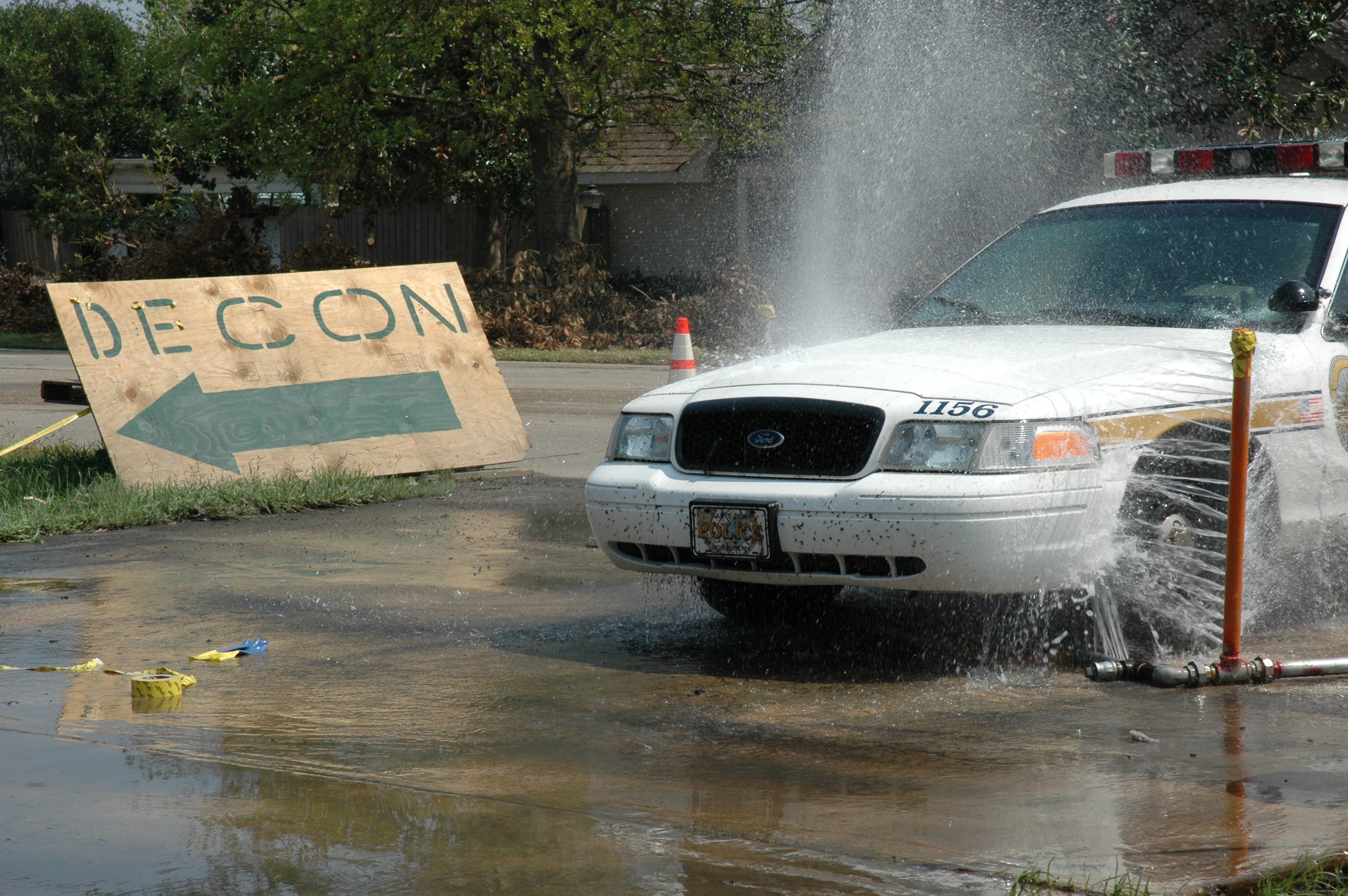 Ein Polizeiauto fährt durch eine Pfütze mit verstreuten Gegenständen, darunter ein Verkehrskegel und eine Tafel mit Text auf dem Boden, während Bäume, Gebäude und ein klarer blauer Himmel im Hintergrund zu sehen sind.