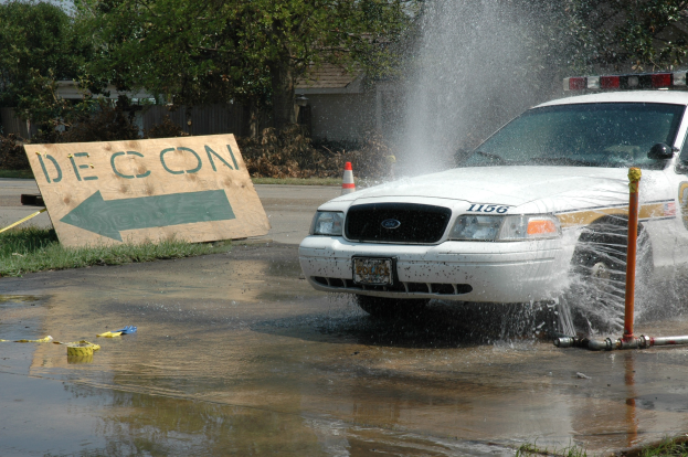 Ein Polizeiauto fährt durch eine Pfütze mit verstreuten Gegenständen, darunter ein Verkehrskegel und eine Tafel mit Text auf dem Boden, während Bäume, Gebäude und ein klarer blauer Himmel im Hintergrund zu sehen sind.