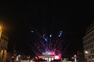 Eine belebte Stadtstraße in Berlin, Deutschland, mit dem Brandenburger Tor im Hintergrund, das von Feuerwerk in den Nachthimmel erleuchtet wird.
