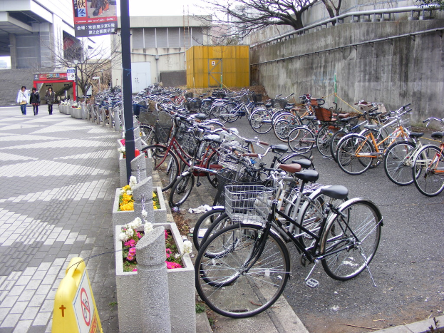 Im Vordergrund fahren Fahrräder auf der Straße nach rechts, während links drei Menschen auf einem Gehweg mit einem Geländer, einer Säule, Blumen und einem Schild gehen; im Hintergrund sind Gebäude, Bäume, ein Unterstand und eine Wand zu sehen, mit einem Banner oben.