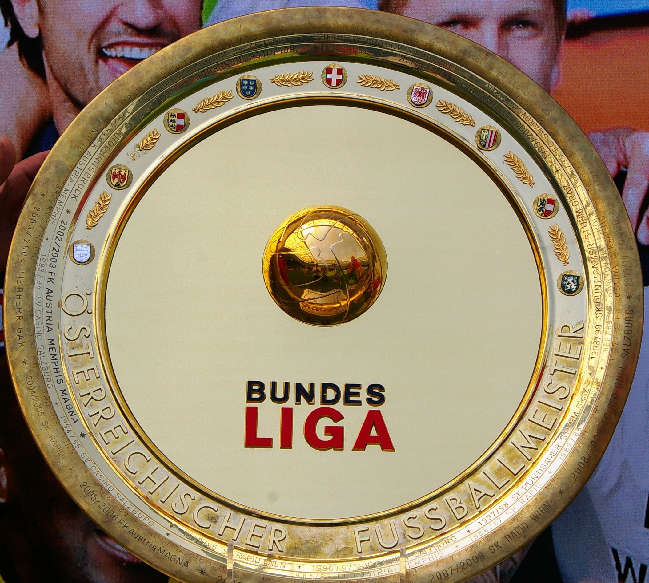 Goldener Schild mit "Bundesliga"-Text in der Hand eines Mannes, drei Personen auf einem Banner im Hintergrund.