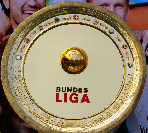Goldener Schild mit "Bundesliga"-Text in der Hand eines Mannes, drei Personen auf einem Banner im Hintergrund.