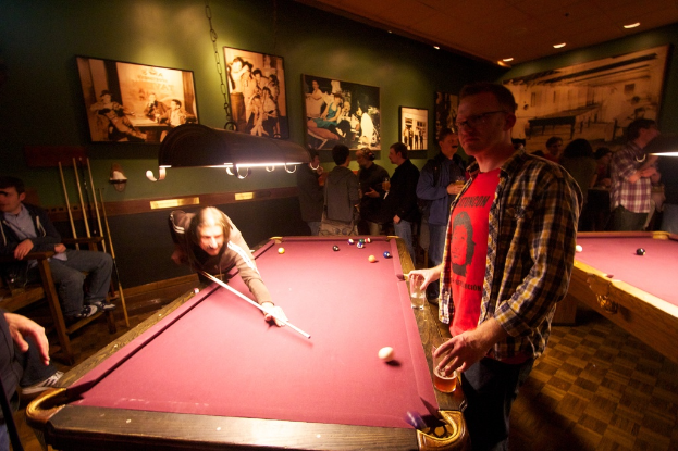 In einem Club spielen zwei Personen Billard an einem von zwei Tischen, während mehrere andere im Hintergrund stehen und sich unterhalten, vor einer cremefarbenen Wand mit ein paar Fotorahmen.
