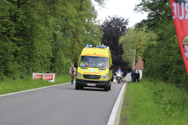 Ambulanz fährt auf Straße mit Fahrradfahrern daneben, umgeben von Gras, Bäumen, Häusern, Strommasten und einem klaren blauen Himmel.