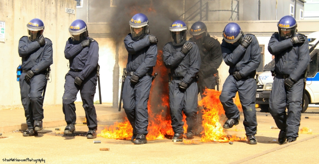 Männer mit Helmen stehen vor einem Feuer mit Gegenständen auf dem Boden, Gebäuden im Hintergrund, einem Fahrzeug, einem Plakat und einer Tafel an der linken Wand und Text unten.