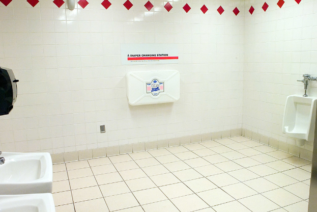 Ein Waschraum mit zwei Waschbecken im Vordergrund, eine Toilettenschüssel rechts und eine Box vor gefliesten Wänden im Hintergrund.