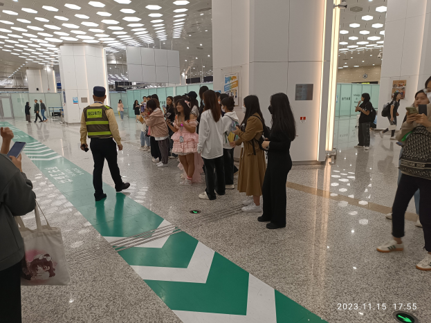 Menschen, die in einer Reihe an einem Flughafen-Sicherheits-Check-in-Schalter stehen und Taschen und Mobiltelefone halten, mit Säulen und Deckenleuchten im Hintergrund.
