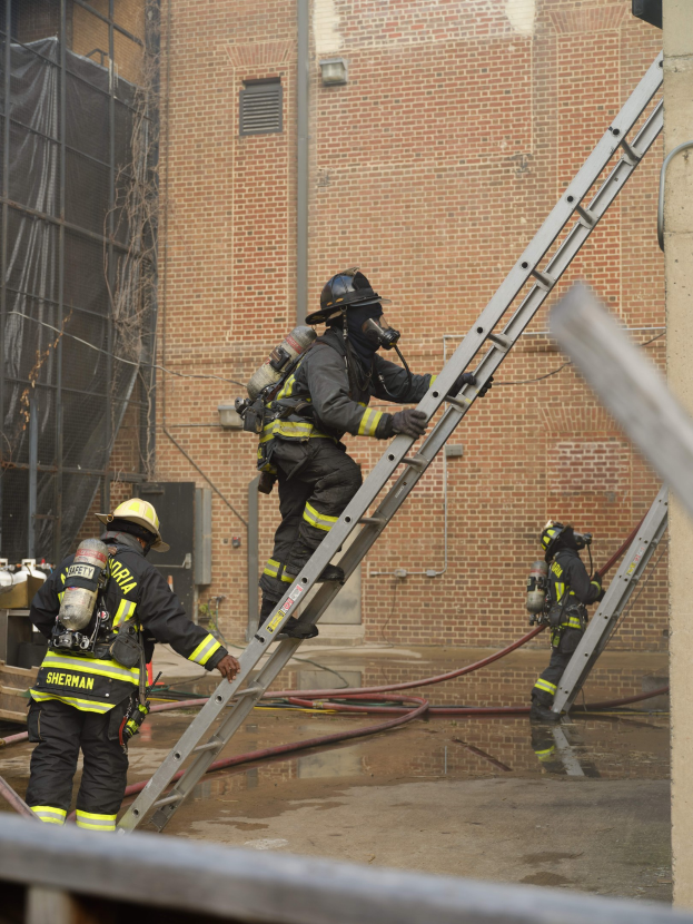 Feuerwehrleute in Helmen und Ausrüstung klettern an einer Leiter vorbei an einem Backsteingebäude mit freiliegenden Rohren und einer Metallstange am Boden, mit einem weiteren Gebäude mit Fenstern und einem Netz im Hintergrund.