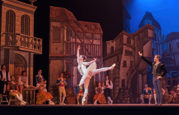 Zwei Tänzer performen auf einer Bühne im Royal Opera House in London, mit einem Publikum im Vordergrund, das auf Stühlen sitzt und steht, und einem Bildschirm im Hintergrund, der Gebäudeabbildungen anzeigt.