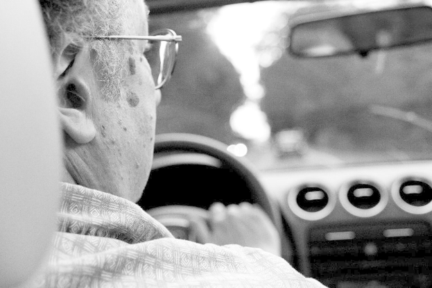 Eine Person mit Brille sitzt auf der linken Seite eines Fahrzeugs, das auf einer Straße mit unscharfem Hintergrund fährt.