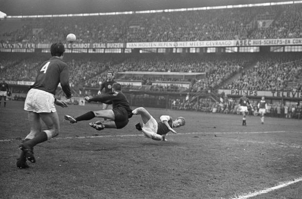 Eine Gruppe von Männern, die auf einem Feld in einem Stadion Fußball spielen, mit Zuschauern auf den Rängen und Bannern im Hintergrund, dargestellt in Schwarz-Weiß.