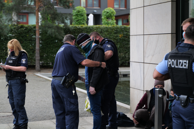Eine Gruppe von Polizeibeamten steht vor einem Gebäude, eine Frau hält ein Buch und einen Stift in der Hand, während mehrere Menschen in der Nähe stehen oder sitzen und Pflanzen, Bäume und Gebäude im Hintergrund zu sehen sind.