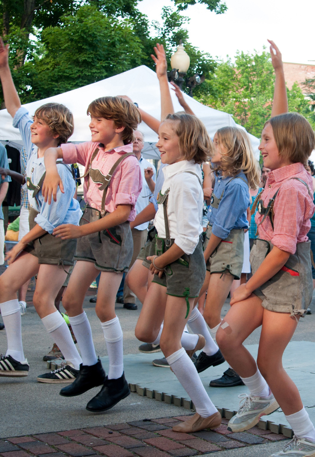 Eine Gruppe von Kindern in traditioneller bayrischer Tracht tanzt auf der Straße, umgeben von einer Menge, Zelten, Laternenmasten, Bäumen, Gebäuden und einem klaren blauen Himmel, wobei einige Kinder Shorts und Schuhe tragen.