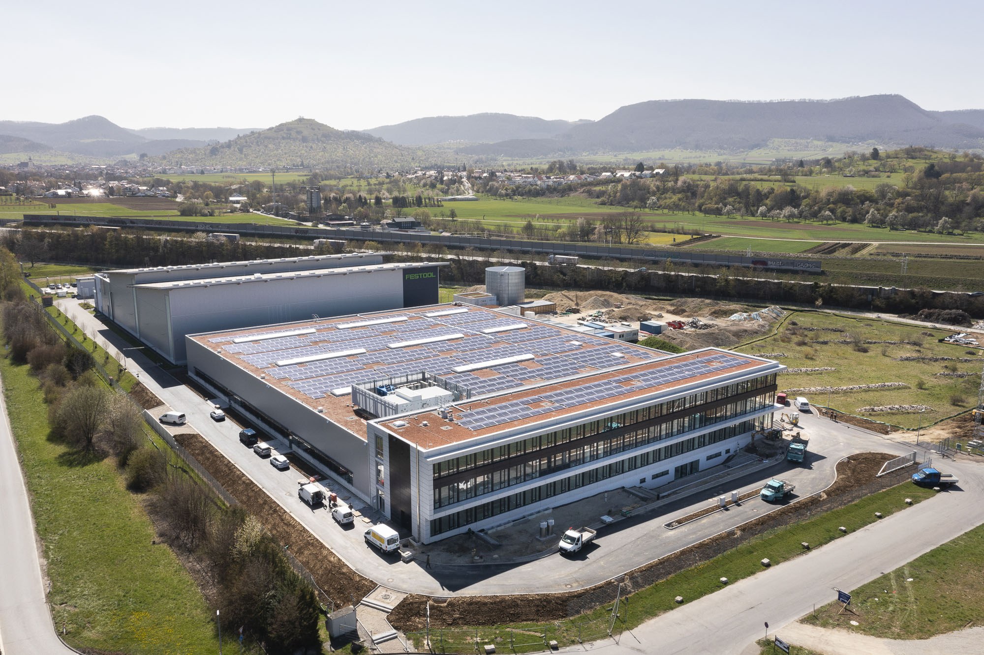 Luftaufnahme eines großen Gebäudes mit Solarpaneelen auf dem Dach, umgeben von Fahrzeugen, Bäumen, Gras und Masten, mit Hügeln und einem klaren blauen Himmel im Hintergrund.