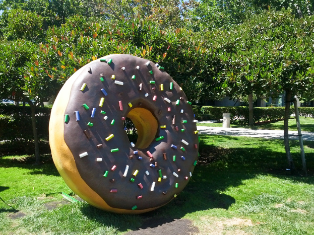 Eine ringförmige Struktur auf Gras mit Bäumen, Pflanzen, einem Poller und Himmel im Hintergrund.