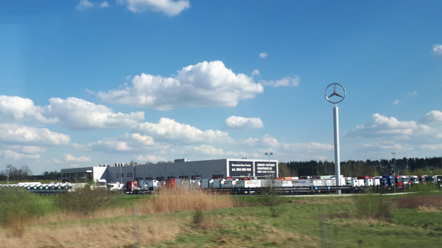 Ein Mercedes-Benz-Werk in Deutschland mit Fahrzeugen auf der Straße, umgeben von Grünflächen und Gebäuden, unter einem bewölkten Himmel.