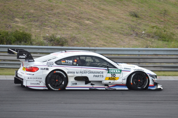 Ein BMW M4 DTM-Rennwagen auf einer Rennstrecke, umgeben von saftig grünem Gras, mit einer Absperrung im Hintergrund.