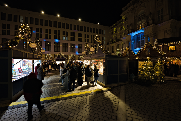 Ein lebendiger Weihnachtsmarkt bei Nacht in einer Stadt, mit Menschen um beleuchtete Stände mit festlicher Dekoration und beleuchtete Fenster im Hintergrund.