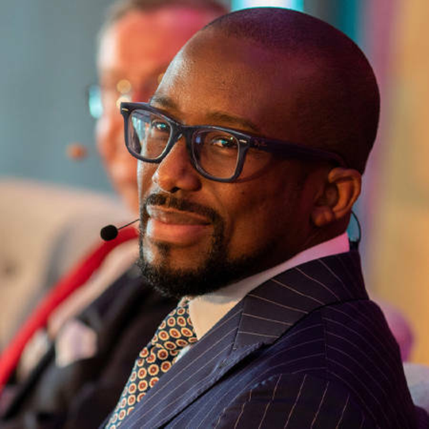 Ein Mann im Anzug und Krawatte sitzt neben einem anderen Mann mit Brille und Mikrofon, mit einem unscharfen Hintergrund.