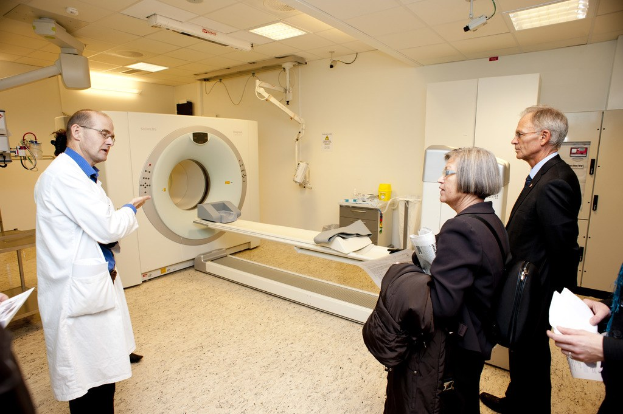 Arzt in weißer Uniform erklärt eine CT-Scan-Maschine zwei Personen in schwarzen Mänteln.